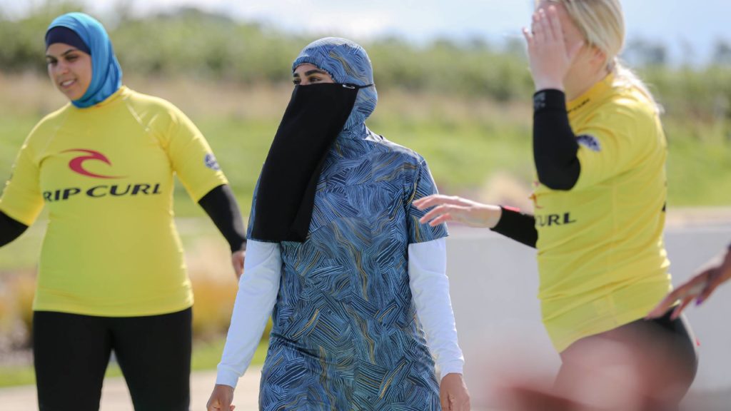 Women in wetsuits follow a surfing demonstration.