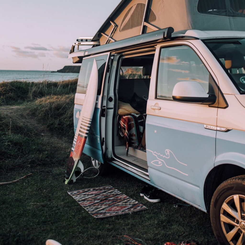 A blue campervan parked by the sea with a surfboard resting against it.